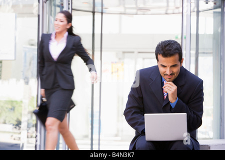 Businessman outdoors in front of building using laptop with businesswoman in background smiling (high key/blur) Stock Photo