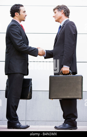 Two businessmen shaking hands outdoors holding briefcases and smiling Stock Photo