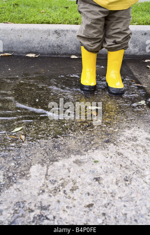 A toddler boy stands in a puddle with yellow rain boots on. Stock Photo
