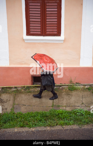 Elderly woman with old umbrella walking on pavement down a street in rain from above. Romania Europe Stock Photo