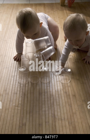 twin baby boys knock over stacked transparent plastic cups on wooden kitchen floor Stock Photo