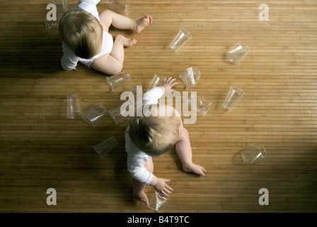 aerial view of twin baby boys playing on bamboo kitchen floor surrounded by scattered transparent plastic cups Stock Photo