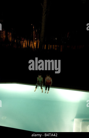 two women looking down sit on the edge of light up pool at night with their feet in water with dark forest in background Stock Photo