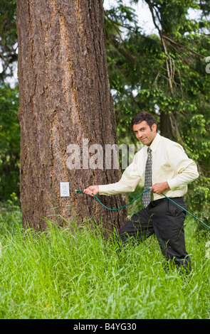 A mixed race businessman holding an electrical extension cord about to plug it in to a electrical outlet on a tree in a grassy f Stock Photo