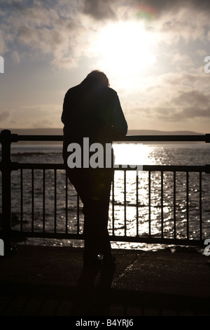 elderly man standing head bowed leaning against a guard rail fence watching the sun set over the sea portstewart Stock Photo