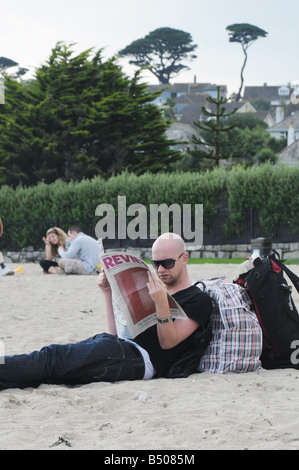 A young man reading the English newspaper The Guardian on a beach in Cornwall, UK Stock Photo