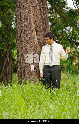 Businessman standing next to a tree holding a extension cord looking down at an electrical outlet on the trunk of a tree in a gr Stock Photo