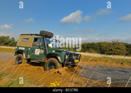 Green Land Rover Defender 90, with high raised air intake and spare wheel on the roof, crossing a flooded forest track. Stock Photo