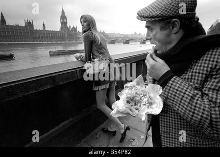 Places London, England. A man eating fish and chips looking at a woman, standing on the bank of the river Thames near the Houses of Parliament. November 1969 Z11367-006 Stock Photo