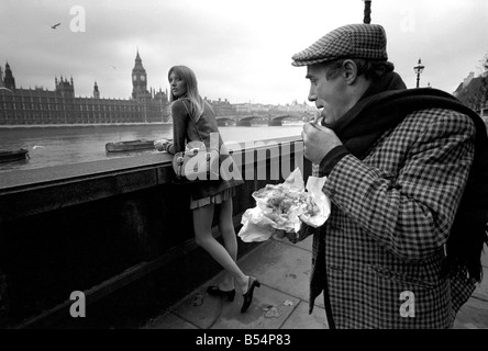 Places London, England. A man eating fish and chips looking at a woman, standing on the bank of the river Thames near the Houses of Parliament. November 1969 Z11367-007 Stock Photo