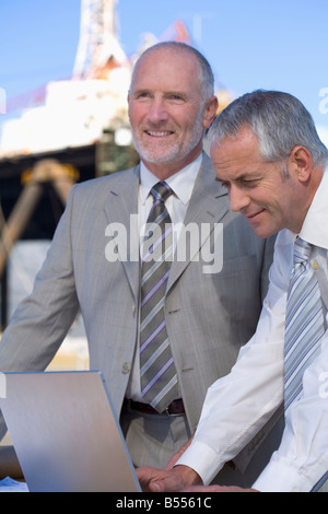 Two engineers studying plans on their laptop, with an oil rig in the background Stock Photo