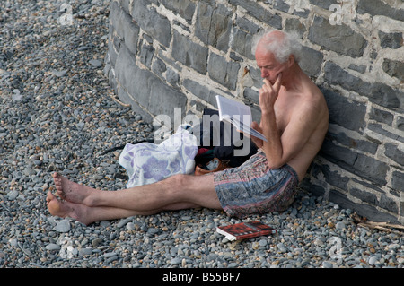 Middle aged man wearing shorts sitting on Aberystwyth beach reading paperback book Wales UK, summer afternoon Stock Photo