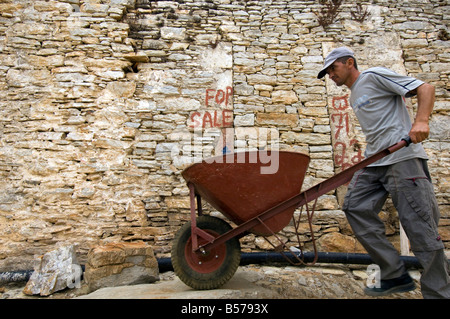 A builder pushes his wheelbarrow past a stone house For Sale in the Old Town, or Chorio, on the Greek Island of Symi Stock Photo