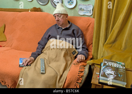 A cold elderly man sleeping on a settee under a blanket wearing a woolly hat Stock Photo