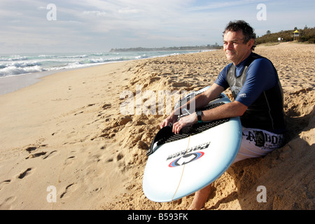 PORTRAIT OF A 50 SOMETHING MALE SITTING ON THE BEACH PREPARING TO GO SURFING HORIZONTAL BDA11151 Stock Photo