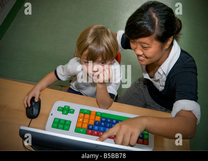 Infant boy 4-5 years in kindergarten working on his colour coded computer keyboard with help from a senior high school teenage girl teacher Stock Photo