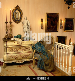 Landing with decoratively painted cream antique chest of drawers and gilt-framed chair with silk throw Stock Photo