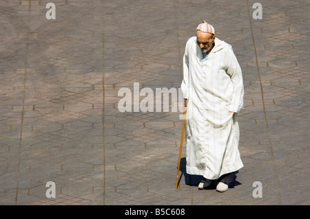 An elderly man with a walking stick and traditional dress walking through the Djemaa El Fna - main market square of Marrakesh. Stock Photo