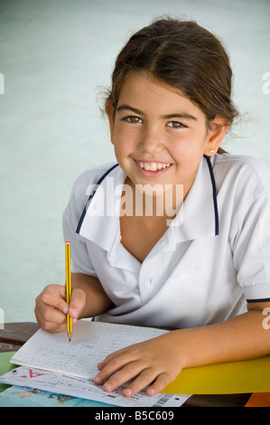 Confident Junior Hispanic schoolgirl smiles to camera whilst catching up with her studies outside in sunny school playground Stock Photo