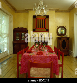 Glass chandelier above red crockery and red cloth on rectangular table in yellow dining room with red picture above fireplace Stock Photo