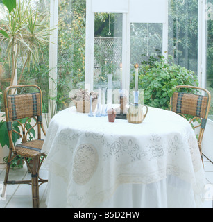 Rattan chairs at circular table with white cloth in small conservatory dining room with view of garden through windows Stock Photo