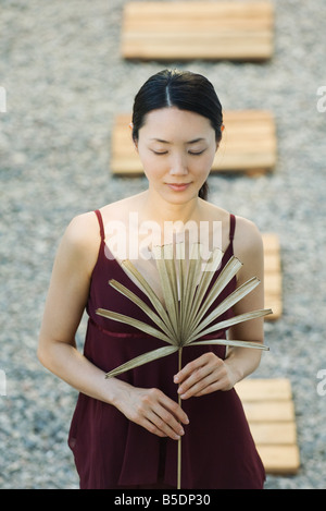 Woman standing in rock garden holding dried palm frond, eyes closed Stock Photo