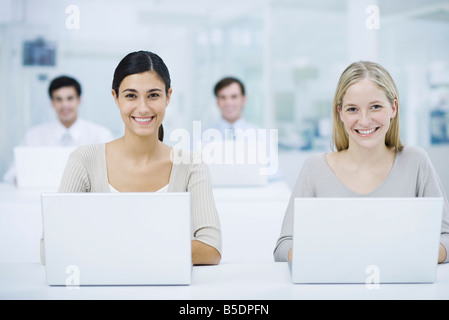 Professional women using laptop computers, smiling at camera, colleagues in background Stock Photo