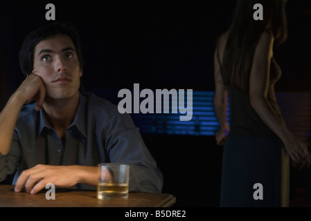 Man sitting near glass of whiskey, looking up, woman dancing in background Stock Photo