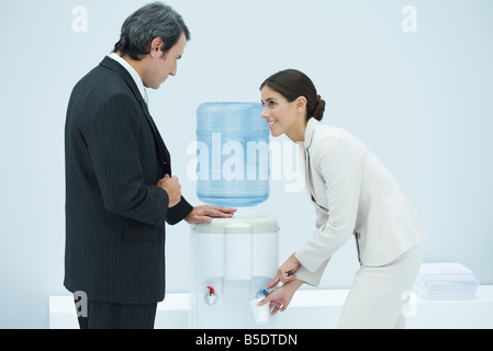 Two professionals chatting beside water cooler, woman filling disposable cup Stock Photo