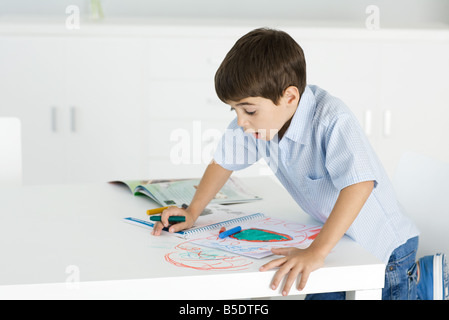 Boy leaning on table, holding crayon, colorful drawing on paper and table Stock Photo