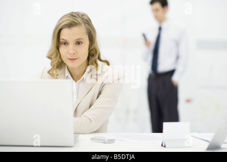Professional woman using laptop computer at desk, man standing in background Stock Photo
