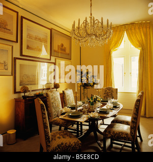 Glass chandelier above oval antique table with upholstered chairs in yellow dining room with pictures and yellow curtains Stock Photo