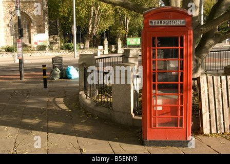 An old style red telephone box stands on a pathway in London on the Thames walkway. An old tree can be seen growing behind Stock Photo
