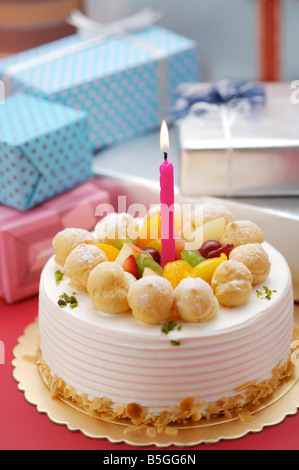 Birthday cake with a lighted candle on top, wrapped presents in the background Stock Photo