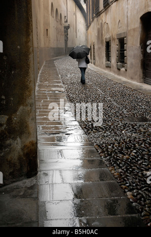 A woman strolls under her umbrella through the wet cobblestone streets of a small Italian mountain town of Bergamo. Stock Photo