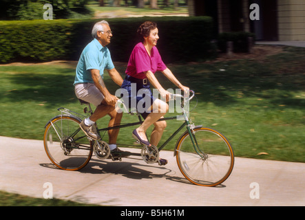Senior Hispanic couple rides tandem bicycle on sidewalk Stock Photo