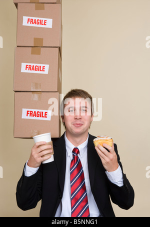 Young businessman holding drink and cake balancing boxes on shoulder Stock Photo