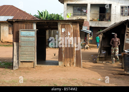 A Nigerian male walks from a falling down lean-to toward a small corrugated shack with a child running in the background Stock Photo