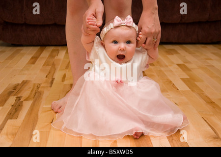 Baby sitting on floor looking surprised with mother holding her hands while learning to walk Stock Photo