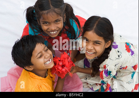 Three indian girl friends sitting holding hibiscus flowers laughing at camera.  Andhra Pradesh, India Stock Photo