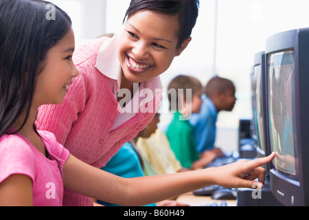 Teacher helping student at computer terminal with students in background (depth of field/high key) Stock Photo