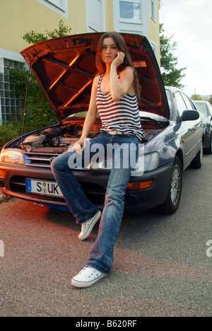 Young woman sitting on her broken down car phoning someone for help Stock Photo