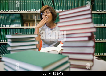 Young, dark-haired woman, a student, sitting at a table laden with piles of books, in front of a bookshelf in a library, yawning Stock Photo