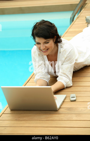 Woman reclined beside a swimming pool, working on a laptop, web surfing, online dating, Internet purchases Stock Photo