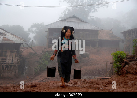 Woman of the Akha Phixo tribe wearing traditional costume carrying two buckets of water in the fog in the village of Ban Mososa Stock Photo