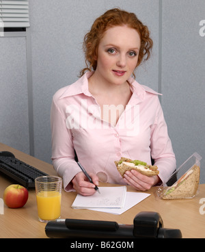 Young woman at her office desk making notes during her lunch break Chicken salad sandwich glass of orange juice apple Stock Photo