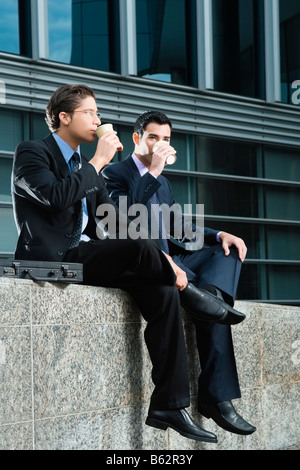 Two businessmen sitting at the edge of a wall and drinking from disposable cups Stock Photo