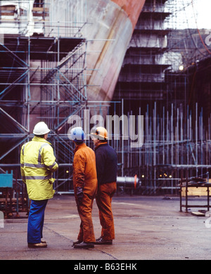 a picture of 3 men working in a dry dock looking at the bow of a ship docked for maintenance Stock Photo