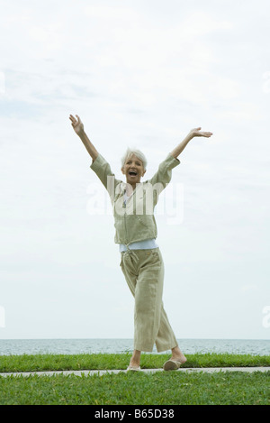 Senior woman standing outdoors in seaside park with arms raised, smiling happily Stock Photo