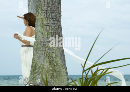 Woman standing beside palm tree, shading eyes with hand, looking at sea, scarf flowing in breeze Stock Photo